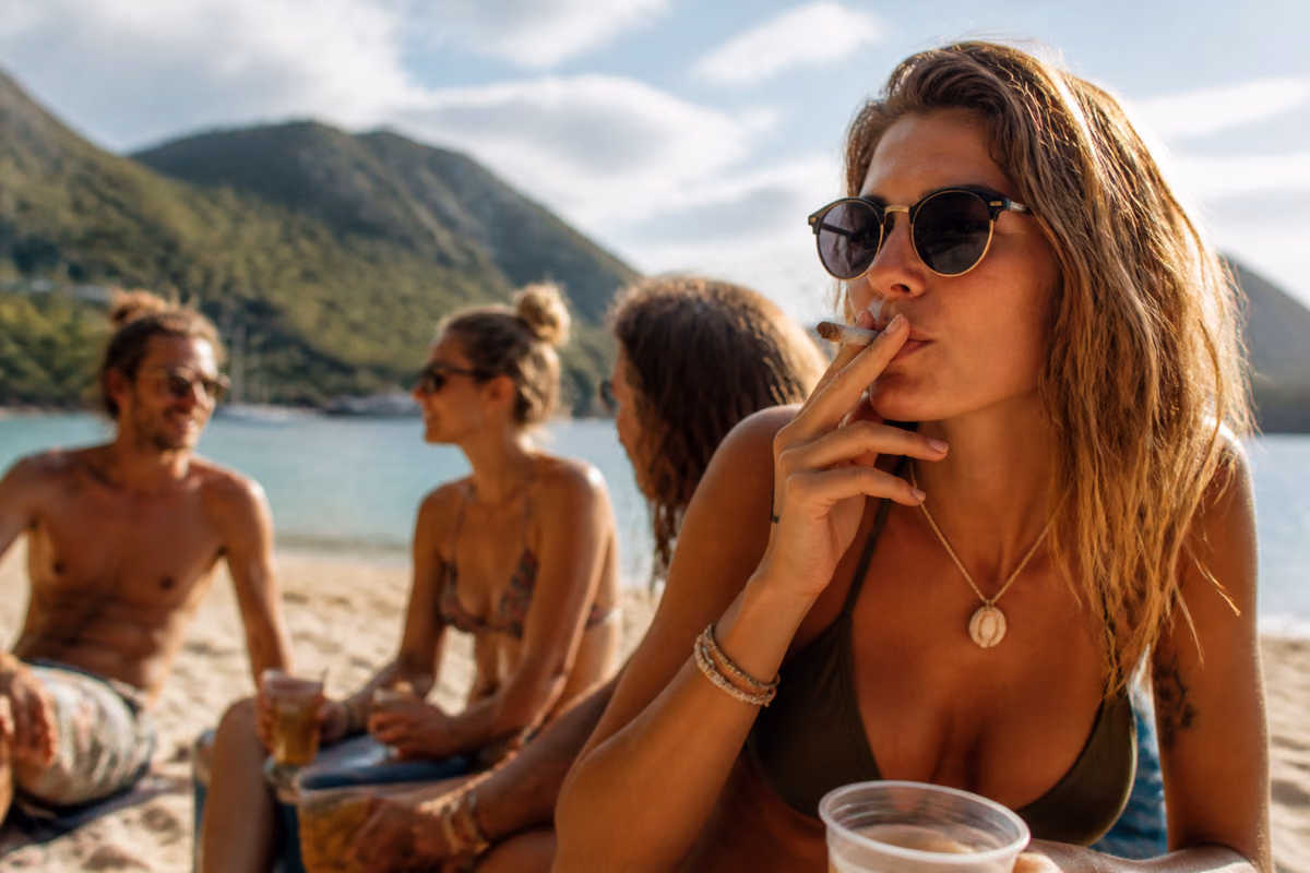 Žena se slunečními brýlemi kouří joint na tropické pláži, v pozadí přátelé s nápoji