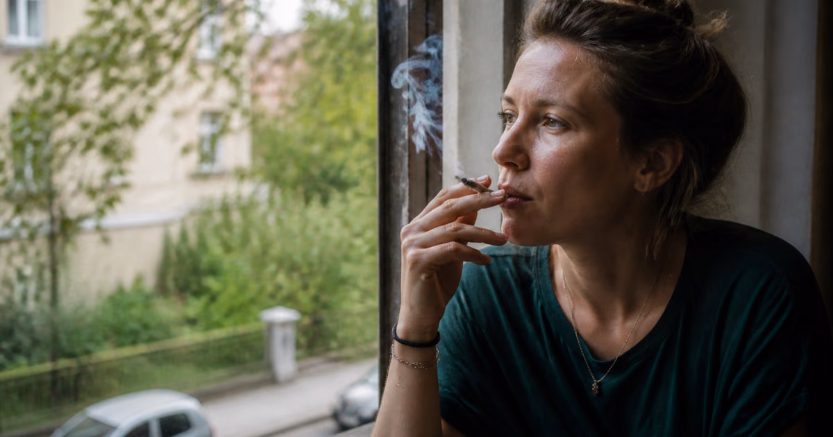 Žena kouří joint u okna a dívá se na ulici plnou zeleně